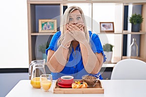 Caucasian plus size woman eating breakfast at home shocked covering mouth with hands for mistake