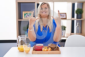 Caucasian plus size woman eating breakfast at home shouting with crazy expression doing rock symbol with hands up