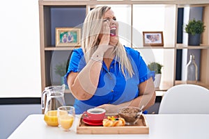 Caucasian plus size woman eating breakfast at home shouting and screaming loud to side with hand on mouth