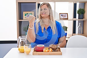 Caucasian plus size woman eating breakfast at home smiling with happy face looking and pointing to the side with thumb up