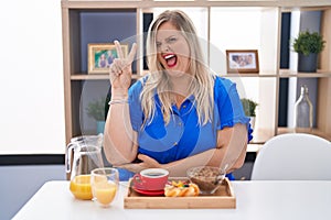 Caucasian plus size woman eating breakfast at home smiling with happy face winking at the camera doing victory sign