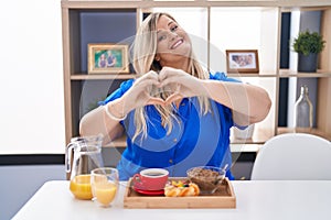 Caucasian plus size woman eating breakfast at home smiling in love doing heart symbol shape with hands