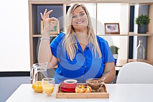 Caucasian plus size woman eating breakfast at home smiling positive doing ok sign with hand and fingers