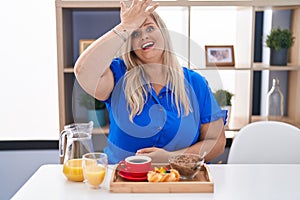 Caucasian plus size woman eating breakfast at home surprised with hand on head for mistake, remember error