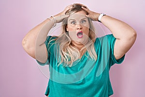 Caucasian plus size woman standing over pink background crazy and scared with hands on head, afraid and surprised of shock with