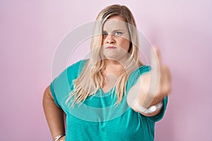 Caucasian plus size woman standing over pink background showing middle finger, impolite and rude fuck off expression