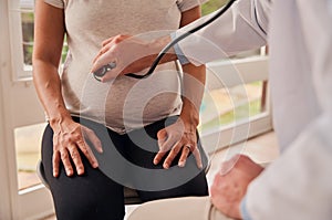 Caucasian pregnant female sitting in clinic while male doctor examines pregnant belly with stethoscope