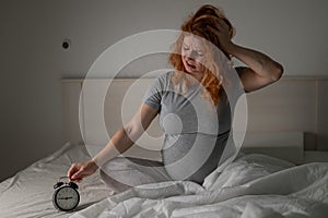 Caucasian pregnant woman sits in bed and looks at the alarm clock. Insomnia.