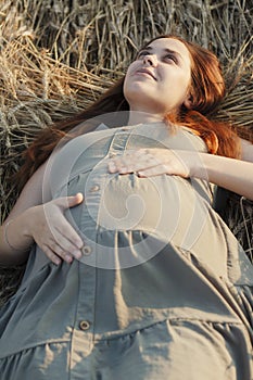 Caucasian pregnant young red-haired woman in dress lying on wheat field in summer at sunset, future mother relaxing in nature,