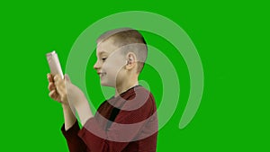 A Caucasian preschool boy on a green background holds a smartphone in front of his face, looks at the screen and smiles.