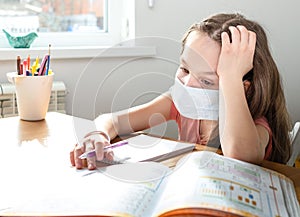 Caucasian preteen girl with medical mask on her face concentrated on her task with schoolbook. Concept of distance learning in