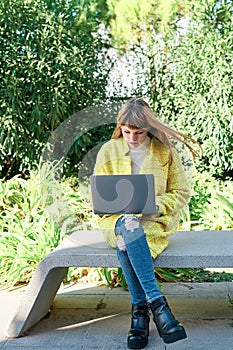 Caucasian pretty young girl, dressed in a yellow jacket, a white sweater, working with the laptop sitting on a park bench