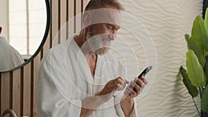 Caucasian relaxed senior old man in bathroom in bathrobe calm mature retired male in bathroom browsing mobile phone