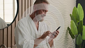 Caucasian relaxed senior old man in bathroom in bathrobe calm mature retired male in bathroom browsing mobile phone