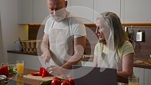 Caucasian retired couple in chefs aprons smiling cooking at home kitchen middle-aged woman wife using laptop culinary