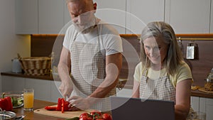 Caucasian retired couple in chefs aprons smiling cooking at home kitchen middle-aged woman wife using laptop culinary