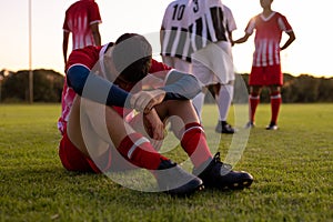 Caucasian sad male athlete sitting on grassy land with team players in background at playground photo