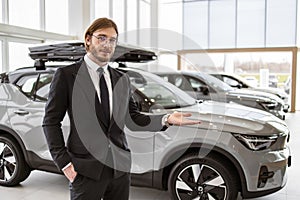 Caucasian salesman looking at camera and pointing at new cars models in showroom