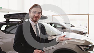 Caucasian salesman looking at camera and pointing at new cars models in showroom