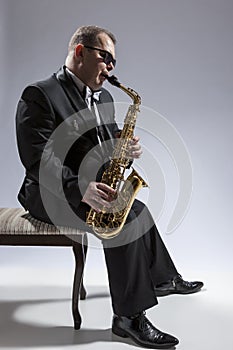 Caucasian Saxophone Player in Sunglasses Playing the Saxophone While Sitting on Chair in Studio Environment.