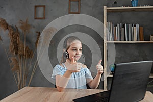 Caucasian schoolgirl sits at home at a laptop and raises her thumb up.