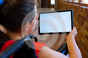 Caucasian schoolgirl sitting in wheelchair and using tablet with copy space at school corridor