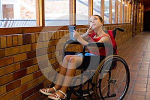 Caucasian schoolgirl sitting in wheelchair and using tablet at school corridor, copy space