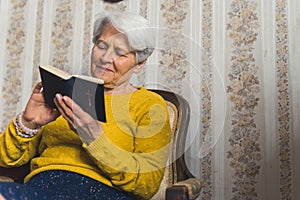 Caucasian senior grandmother in her 70s sitting on an old-fashioned chair and peacefully reading the Bible. Spirituality