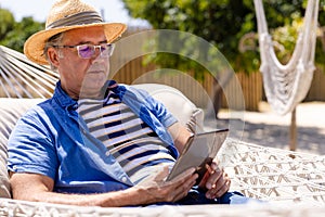 Caucasian senior man wearing hat using digital tablet while resting on hammock at beach