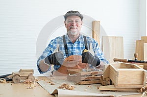 Caucasian senior old bearded man carpenter in apron, hat and gloves sitting smiling, thumbs up, working in workshop, tools machine