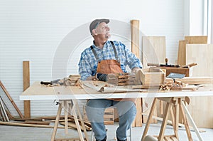 Caucasian senior old white bearded man carpenter in apron, hat and gloves working in workshop, thinking looking out of window,