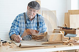 Caucasian senior old white bearded man carpenter in apron, hat and gloves working in workshop, use pencil sketching a design on