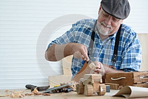 Caucasian senior old white bearded man carpenter in apron and hat using chisel working in workshop, tools machine small wooden toy