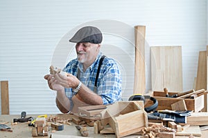 Caucasian senior old white bearded man carpenter in apron and hat working in workshop, holding and looking at handmade wood toy,