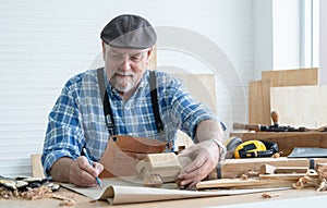 Caucasian senior old white bearded man carpenter in apron and hat working in workshop, sketching a car model design on paper,
