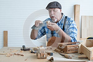 Caucasian senior old white bearded man carpenter in apron and hat working in workshop, use sandpaper polishing and blowing dust