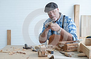 Caucasian senior old white bearded man carpenter in apron and hat working in workshop, use sandpaper polishing handmade wood toy,