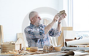 Caucasian senior old white bearded man carpenter working in workshop, holding, looking at handmade wood car toy in detail, tools