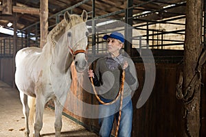 Senior woman horse breeder standing with white horse in barn