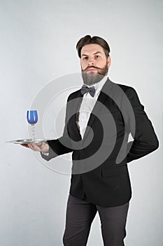 Caucasian servant with a beard in a formal business suit stands with a steel tray in his hand and a glass of wine on a white solid