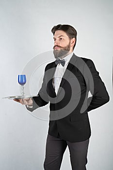 Caucasian servant with a beard in a formal business suit stands with a steel tray in his hand and a glass of wine on a white solid