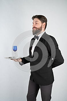 Caucasian servant with a beard in a formal business suit stands with a steel tray in his hand and a glass of wine on a white solid