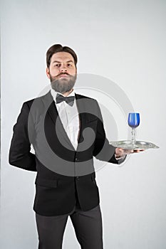 Caucasian servant with a beard in a formal business suit stands with a steel tray in his hand and a glass of wine on a white solid