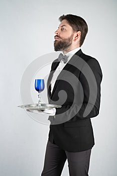 Caucasian servant with a beard in a formal business suit stands with a steel tray in his hand and a glass of wine on a white solid