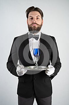 Caucasian servant with a beard in a formal business suit stands with a steel tray in his hand and a glass of wine on a white solid