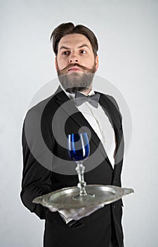 Caucasian servant with a beard in a formal business suit stands with a steel tray in his hand and a glass of wine on a white solid