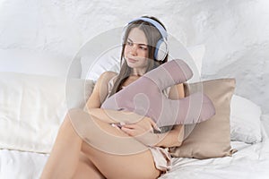 Caucasian sexy girl with headphones listens to music while sitting in bed in silk pajamas