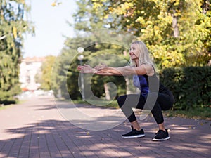 Caucasian smiling middle-aged athletic woman doing deep squat on footway in urban park, outdoor, selective focus photo