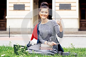 Caucasian student girl shows thumb up on campus lawn with books.