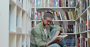 Caucasian student man reading book, sitting among stack of books on library floor, side view.